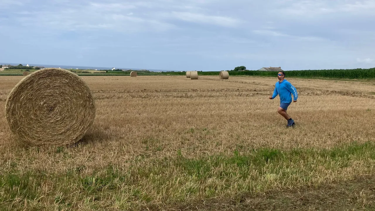 Yann running across a harvested field in Brittany, round straw bales behind him and the sea on the horizon.